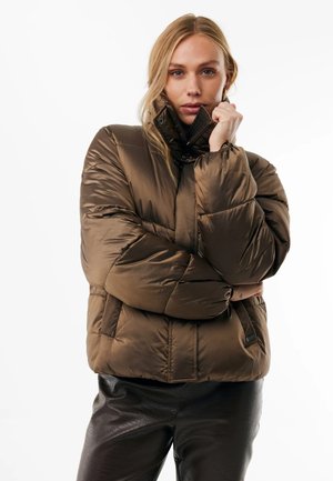Frau mit blondem Haar, die eine braune gesteppte Jacke trägt, die zugezippt ist, und schwarze strukturierte Hosen, posiert mit der Hand nahe am Gesicht vor einem weißen Hintergrund.