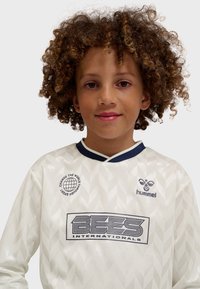 Child with curly hair wearing a white sports jersey with "BEES Internationals" and "hummel" logos facing forward against a plain background.