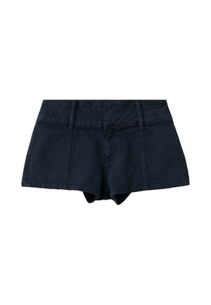 Femme debout devant un fond bleu pâle, portant un haut dos nu blanc, un short noir et des sandales marron cloutées, les mains derrière le dos.