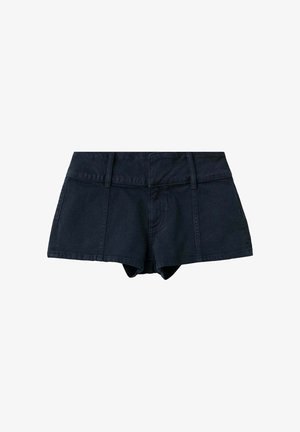 Femme debout devant un fond bleu pâle, portant un haut dos nu blanc, un short noir et des sandales marron cloutées, les mains derrière le dos.