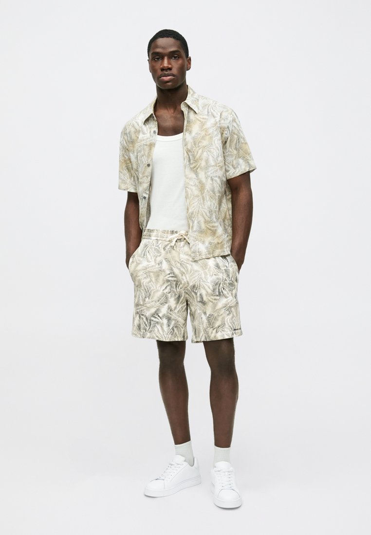 Chemise beige à manches courtes avec motif de feuilles de palmier, assortie à un short coordonné. Débardeur blanc en dessous, baskets blanches, coupe décontractée.