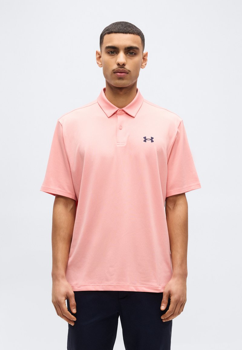 Νέος άνδρας που φοράει ένα ανοιχτό ροζ πόλο πουκάμισο Under Armour και σκοτεινό παντελόνι, στέκεται μπροστά σε μια απλή ανοιχτογκρί φόντο.