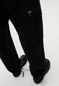 Osoba v černých cargo kalhotách s boční kapsou označenou "PULL&BEAR" a černými šněrovacími botami na světlém pozadí.