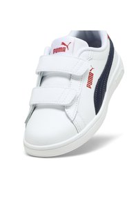 Baskets blanches avec deux bandes Velcro, accent marine sur le côté, logo sur la languette et une texture de surface lisse. Semelle en caoutchouc flexible.