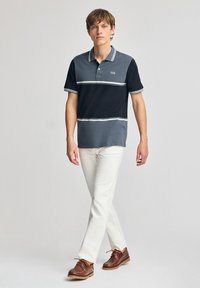 Pikétröja i svart, grått och ljusgrått med horisontella ränder. Har krage och knapplist. Bärs med vita byxor och bruna skor.