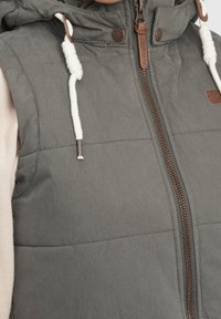 Grau gepolsterte Weste mit Kapuze, ausgestattet mit einem Frontreißverschluss und Zugbandsdetails. Das Material hat eine glatte Textur. Akzente sind braune Beschläge.