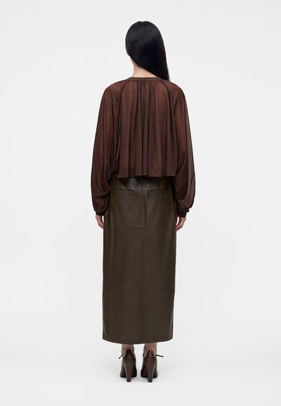 Personne aux longs cheveux foncés se tient de dos, portant un chemisier marron transparent, une jupe en cuir marron foncé et des chaussures marron à talons.
