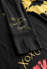 Must mustriline särgis, millel on kuldse trükiga tekst varrukal: "Enesearmastus, Jaga armastust, Korda." Pehme puuvillane kangas sileda tekstuuriga.