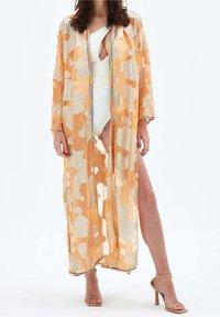 Žena v bílých plavkách a dlouhém průhledném oranžovém kimonu s květinovým vzorem a vícebarevnými lemy, doplněná o nahé vysoké podpatky.