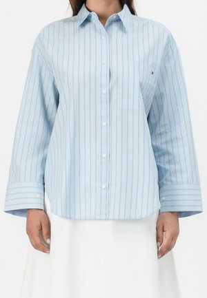 Camisa azul claro abotonada con finas rayas verticales oscuras y puños anchos, usada por una persona con falda blanca.