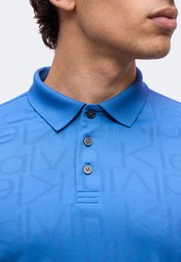 Buste masculin portant un polo bleu Calvin Klein avec un col et trois boutons noirs, tourné vers la droite, sur un fond clair uni.