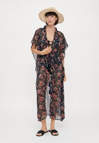 Gjennomsiktig svart kimono med fargerikt blomsterdesign, knyting foran, brede ermer og ankel-lengde. Kombinert med svarte sandaler og stråhatt.