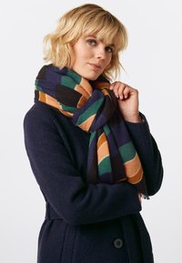 Wollschal mit geometrischem Muster in Orange, Grün und Marineblau, um den Hals gewickelt, mit Fransenenden. Strukturierte Oberfläche.