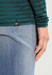 Grön och mörkt randig långärmad skjorta med en svart triangulär etikett, bärs över ljusblå denimjeans. Tyget verkar mjukt och texturerat.