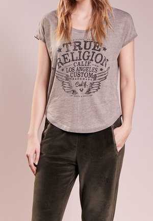 Graues T-Shirt mit kurzen Ärmeln, bedruckt mit einem schwarzen Grafikmotiv mit Flügeln und Text, kombiniert mit dunkelgrünen Hosen aus weichem Stoff mit Taschen.