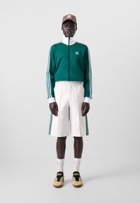 Grön jacka med dragkedja, vit krage och Adidas-logotyp, krämfärgade plisserade shorts med gröna sidsidoränder, vita strumpor och ljusbruna sneakers.