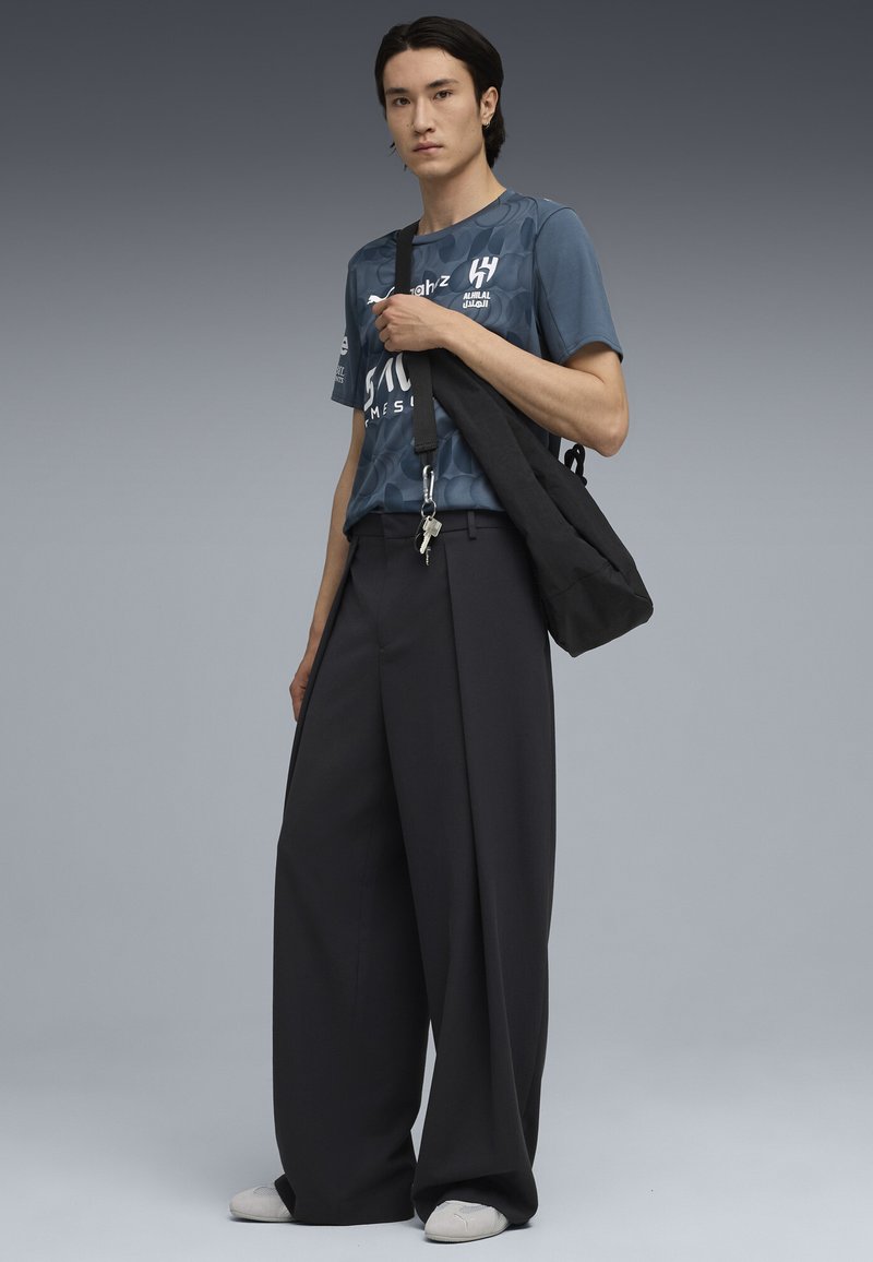 Camiseta gris con estampado, pantalones negros de corte amplio y un bolso negro con llaves adjuntas. Calzado blanco completa el conjunto.