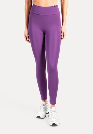 Lila Leggings mit hoher Taille aus glattem, dehnbarem Stoff. Kombiniert mit weißen Sportschuhen und betont einer figurbetonten Silhouette.