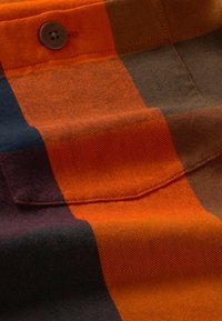 Chemise en flanelle avec un grand motif à carreaux orange, marine, marron et rouge foncé, décoration par boutons, et tissu doux et texturé.