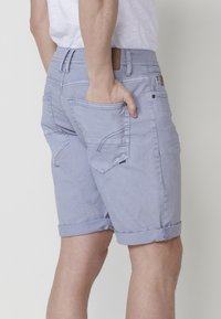 Vīrietis valkā gaiši zilu džinsu šortu ar rullētām malām, roku pielicis aizmugures kabatē, un balta krekla pret vienkāršu fona.