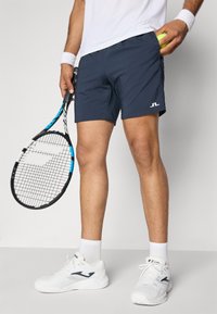 Calções de treino azul-marinho com um logótipo branco, combinados com ténis brancos e uma raquete de ténis azul. Uma bola de ténis amarela está no bolso.
