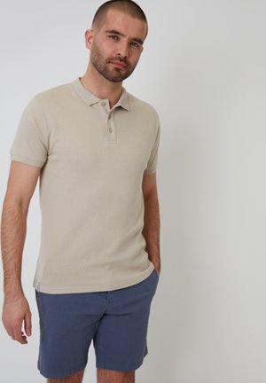 Camisa polo beige con un patrón texturizado sutil, mangas cortas y una tapeta de tres botones. Combinada con pantalones cortos azul marino.