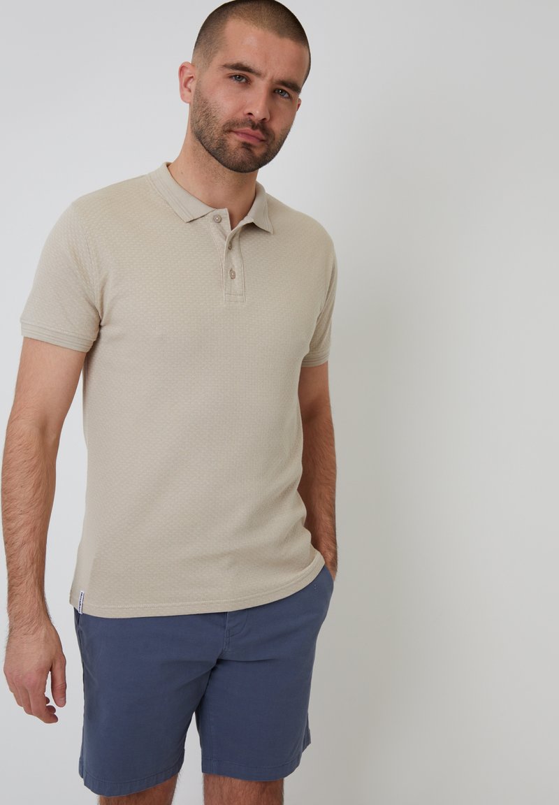 Beige polo shirt met een subtiele gemêleerde structuur, korte mouwen en een drieknoopsluiting. Gecombineerd met marineblauwe shorts.