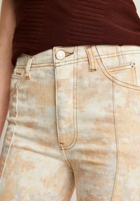Jeans de talle alto en un color crema desgastado con patrones de salpicaduras en naranja y beige. Presenta costuras visibles y un cierre frontal con botón.