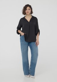 Blusa nera, ampia e comoda, con colletto e tessuto a trama, abbinata a jeans a gamba larga blu chiaro e scarpe con accenti argentati.