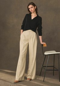 Sort V-udskæring sweater med ribbet tekstur parret med højtaljede, plisserede beige bukser. Modellen står ved siden af en minimalistisk sort stol.