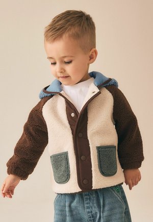 Ung dreng, der kigger ned, iført en hvid og brun fleecejakke med blåt hætte og grå lommer samt blå jeans mod en beige baggrund.
