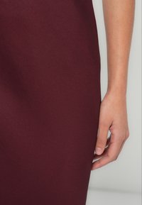 Robe ajustée bordeaux à la texture lisse, mettant en avant un gros plan d'une main reposant sur le tissu, soulignant la douceur du matériau.