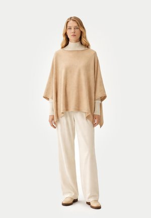 Femme debout portant un poncho beige sur un col roulé crème avec un pantalon large crème et des chaussures beige sur un fond uni.
