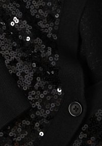 Schwarze Strickjacke mit Lurex auf einer Seite, glatter Textur und einem runden schwarzen Knopf auf der gegenüberliegenden Seite.