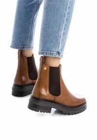 Stivali ankle in pelle marrone con pannelli laterali in rete nera e una suola spessa e testurizzata. Punta arrotondata e piccolo dettaglio metallico.