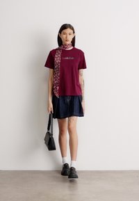 Burgunderfarget bomull T-skjorte med logo, sammen med et denimskjørt. Tilbehør inkluderer et mønstrete rosa skjerf og svarte sko.