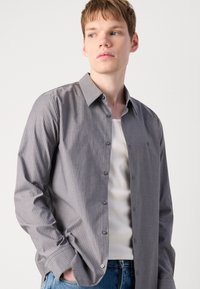 Camisa de botones a rayas en tonos grises, con cuello, de mangas largas, fabricada en un tejido ligero, con un logo sutil en el pecho, combinada con una camiseta sin mangas blanca.