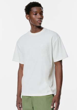 T-shirt in cotone bianco con collo rotondo, maniche corte e un logo goffrato discreto sul lato sinistro del petto. Vestibilità ampia.