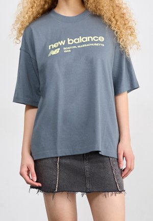 Person iført en blå oversized New Balance T-shirt med gul logo og sort frynset denim mininederdel, stående mod en hvid baggrund.