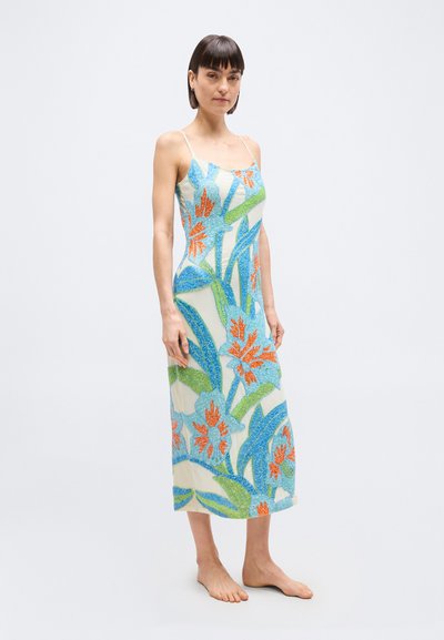 Γυναίκα που στέκεται ξυπόλυτη φορώντας αμάνικο φόρεμα μήκους μέχρι τους  αστραγάλους με μεγάλα μπλε, πράσινα και πορτοκαλί λουλουδένια σχέδια σε ανοιχτό φόντο.