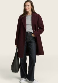 Bordeauxfarbener Wollmantel mit Reverskragen und doppelt verdeckten Knöpfen, kombiniert mit einem grauen Pullover, dunklen Jeans und weißen Sneakers. Schwarze Handtasche.