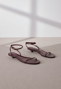 Flache Sandalen aus burgunderrotem Leder mit zwei dünnen Riemen und einem verstellbaren Knöchelriemen, ausgestattet mit einem kleinen Absatz und einer glatten Oberfläche.