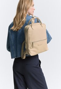 Beige Leder-Rucksack mit zwei oberen Griffen, verstellbarem Schultergurt, vorderer Reißverschlusstasche und glatter Oberfläche. Über einer Jeansjacke getragen.