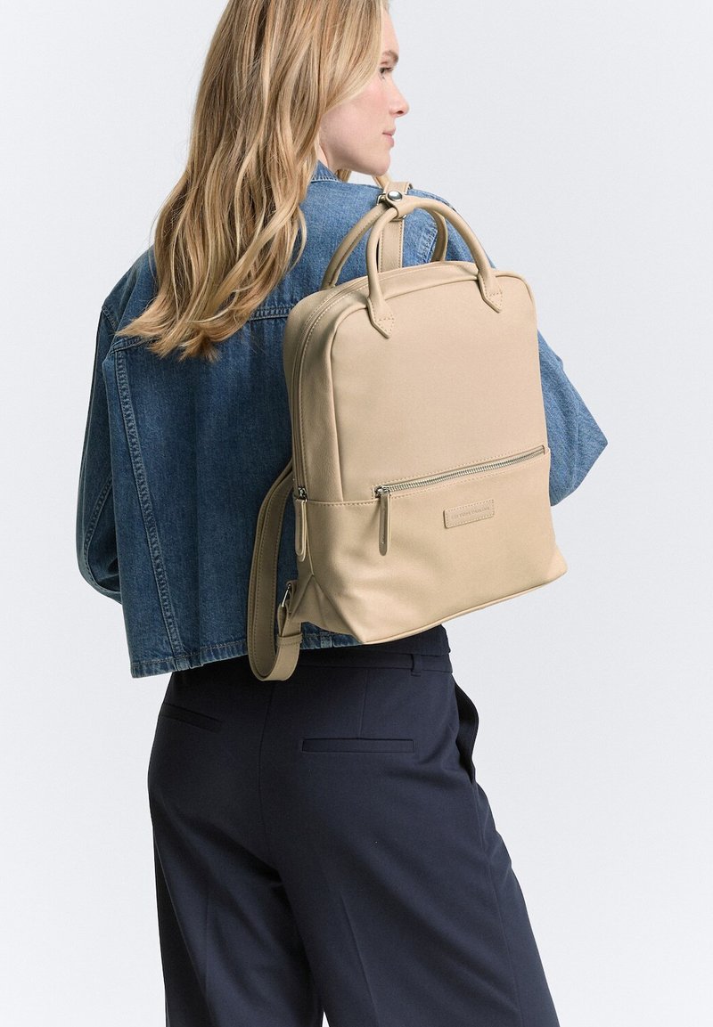 Beige Leder-Rucksack mit zwei oberen Griffen, verstellbarem Schultergurt, vorderer Reißverschlusstasche und glatter Oberfläche. Über einer Jeansjacke getragen.