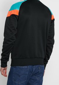 Sudadera negra con detalles en turquesa y naranja en los hombros, textura suave, puños y dobladillo acanalados, y corte holgado. Hombros ligeramente acolchados.