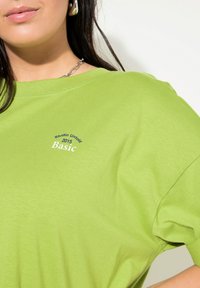 Grøn bomulds T-shirt med rund hals, der har et lille trykt logo, som inkluderer "Studio Unfold 2015 Basic" i kontrastfarver.