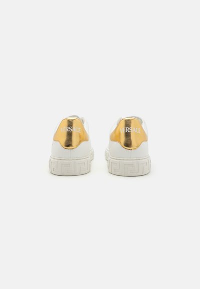 White sneakers featuring a shiny gold heel accent with "VERSACE" branding. Textured rubber sole with a geometric pattern. Rounded design.