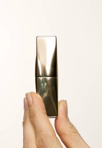 Gull metallisk leppestift rør med et elegant rektangulært design, jevn tekstur og reflekterende overflate, holdt mellom fingrene mot en lys bakgrunn.