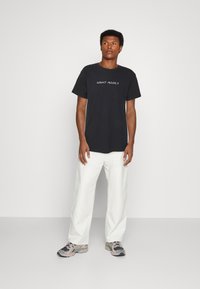 Schwarzes Baumwoll-T-Shirt mit dem Text "NIGHT ADDICT" in Weiß, kombiniert mit lockeren weißen Hosen und mehrfarbigen Sneakers. Schlichter Hintergrund.