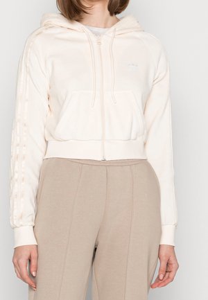 Femme portant un sweat à capuche crème avec zip et un pantalon beige assorti, debout devant un fond uni clair.